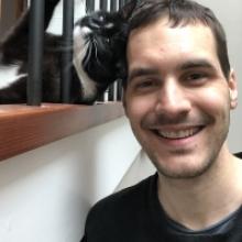 A photo of Andrew and his cat. Andrew is a white, normal looking man. His cat is a black and white tuxedo, domestic short hair. The cat looks disgruntled.