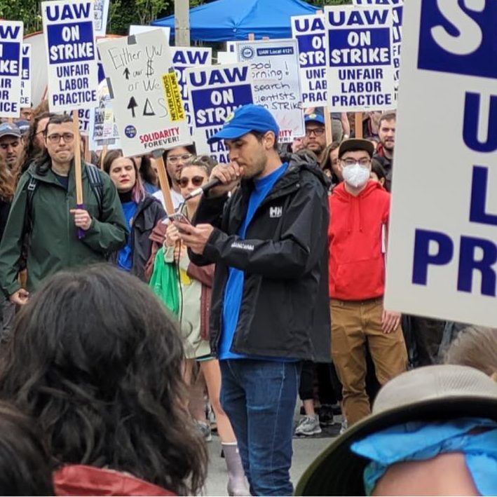 a man wearing a blue hat holding a microphone is shown speaking to a crowd holding up signs saying that UAW is on strike